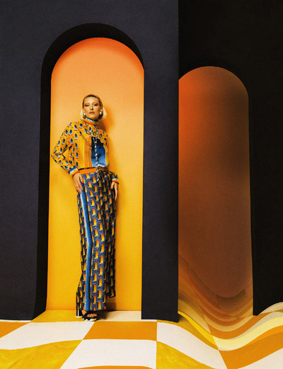 Maggie wears white and gold resin dome earrings, printed silk scarf blouse, arch landscape beaded bolo tie, tan monogram logo leather belt, printed silk trousers, and navy and white patent leather heeled sandals, all by Casablanca. - © System Magazine